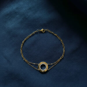 An 18k gold crystal circle bracelet with a double chain design resting on a dark blue textured cloth background.