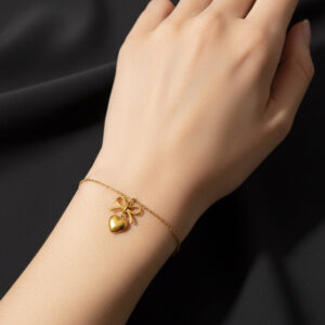 A close-up view of a woman wearing a delicate gold heart bow bracelet on her wrist against a clean black background.