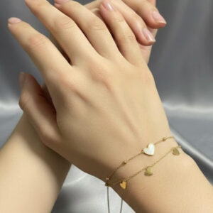 A premium gold double strand heart bracelet featuring a central white heart motif and trailing gold heart charms displayed on a woman's wrist.