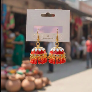 A pair of premium gold red jhumka earrings with red enamel coating and kundan stones held up in an outdoor marketplace setting.
